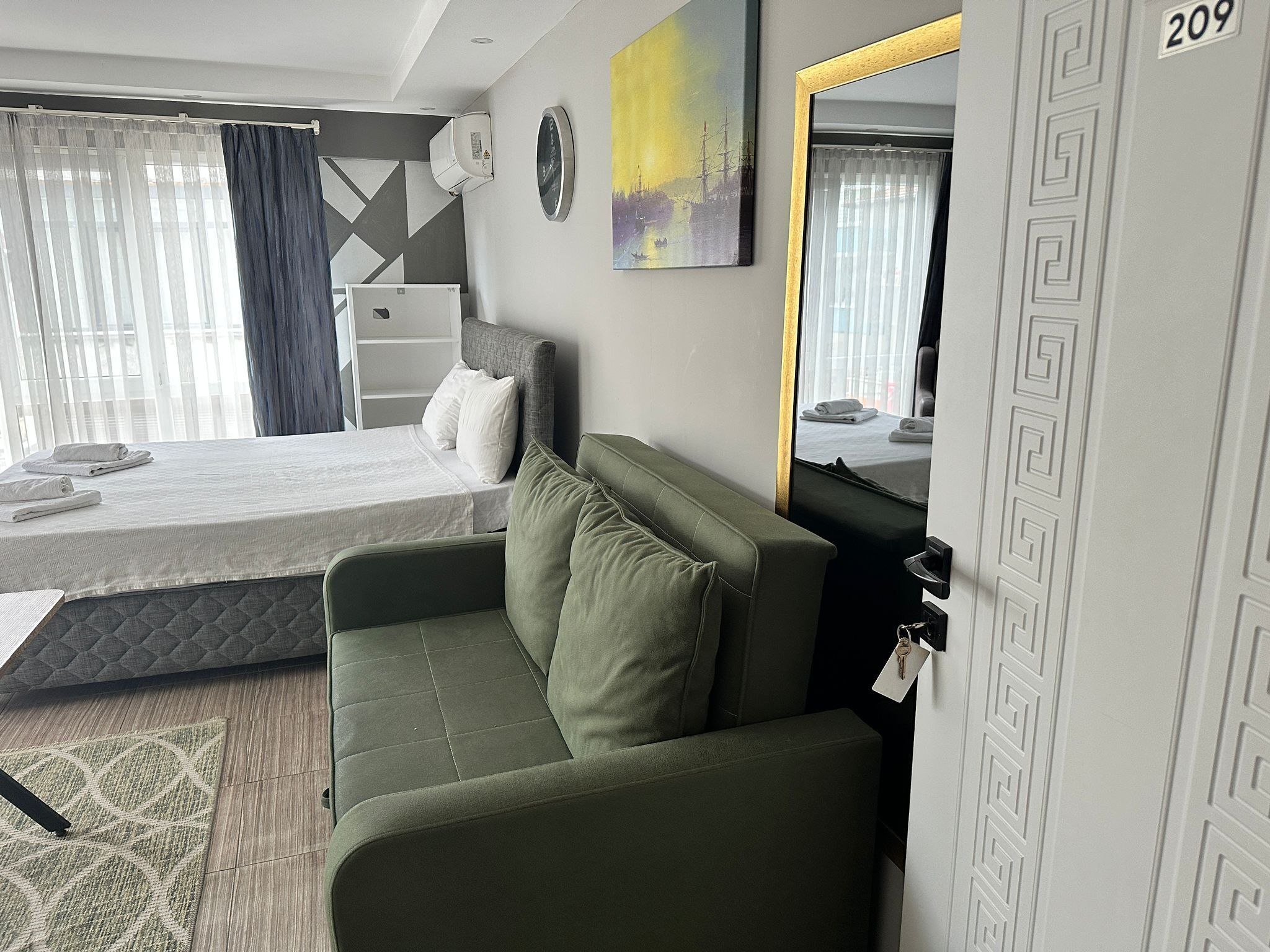 A modern hotel room with a bed, a green sofa, a large mirror, and a view of the city through the window.