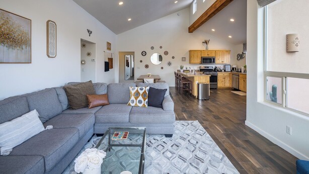 A spacious living room with a gray sectional sofa, a glass coffee table, and a patterned rug. A kitchen with wooden cabinets and a central island is visible in the background. - Slickrock Sanctuary (Moab)
