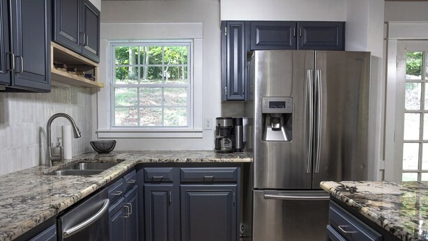 A modern kitchen with dark cabinets, a stainless steel refrigerator, a built-in coffee maker, and a window with a view of trees.