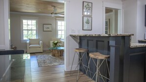 A modern kitchen with dark cabinetry, a marble countertop, and two bar stools. There is a ceiling fan and a window with a view of greenery.
