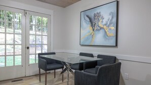 A dining area with a glass table and six chairs, a large abstract painting on the wall, and a door leading to an outdoor area.