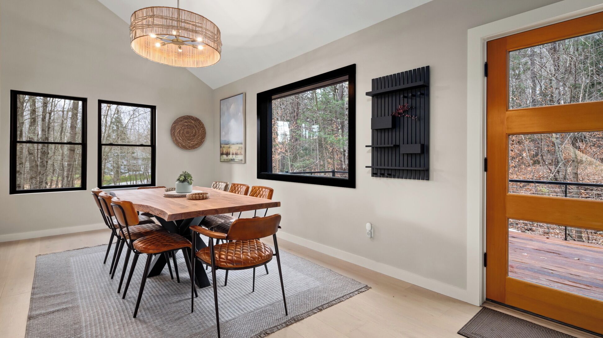 A dining area with a wooden table and chairs, large windows with forest views, a woven wall hanging, and a modern light fixture.
