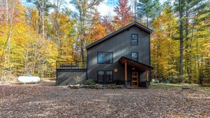 A modern two-story cabin with a dark exterior, surrounded by a forest with autumn foliage.