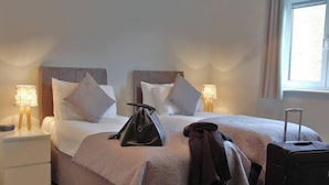 Une chambre d’hôtel avec un lit, deux tables de chevet, une lampe, une valise et une fenêtre.