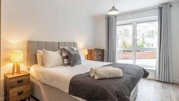 A modern bedroom with a large bed, a bedside table with a lamp, and a view of the outdoors through a large window.