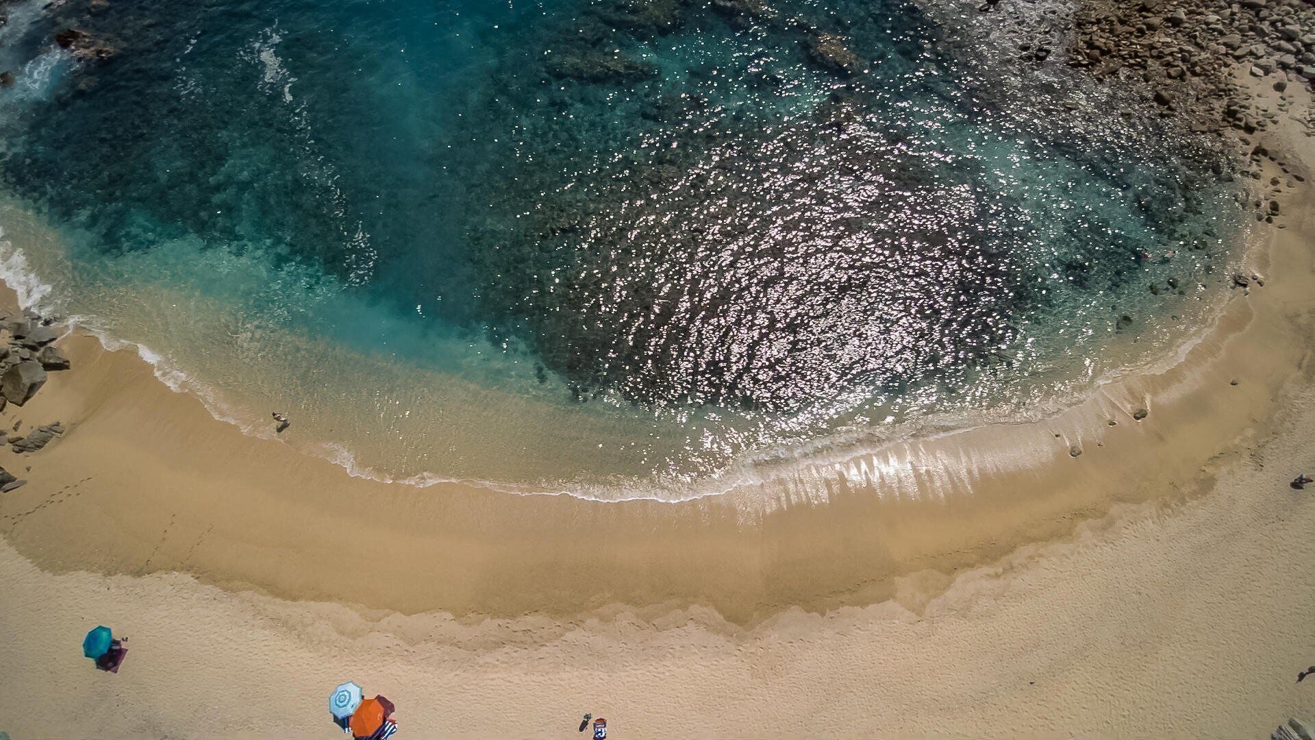 An aerial view of a beach with clear turquoise waters, sandy shore, and a few scattered rocks.