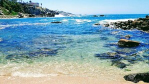 Une plage à l’eau d’un bleu limpide, avec une côte rocheuse et un bâtiment situé au loin, sur une colline.