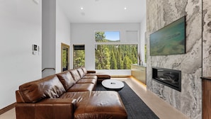 A modern living room with a leather sectional sofa, a round coffee table, a fireplace, and a large window offering a forest view. - Iwtbk4 (Keystone)