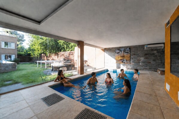 An indoor swimming pool with a clear view of the surrounding area, including a patio and a building with a stone facade. - Buenos Aires de Ezeiza Quinta Vacacional - Dos Departamentos (La Unión)