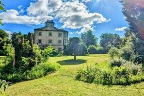 Een groot, historisch gebouw met een symmetrische gevel, omgeven door weelderige begroeiing en een verzorgd gazon.