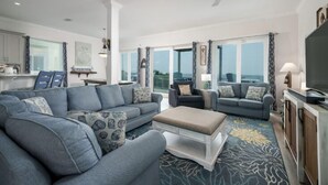 Ein geräumiges Wohnzimmer mit einer großen Eckcouch, einer kleineren Couch und einem Couchtisch. Ein Fernseher steht auf einer Holzkommode, und das Zimmer verfügt über große Fenster mit Vorhängen.