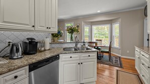 A kitchen with white cabinets, granite countertops, and a dishwasher. There is a dining area with a table and chairs, and a window with a view of greenery. - Near Downtown Cary and Raleigh - 5 room King beds, walking distance to Bond Park (Cary)