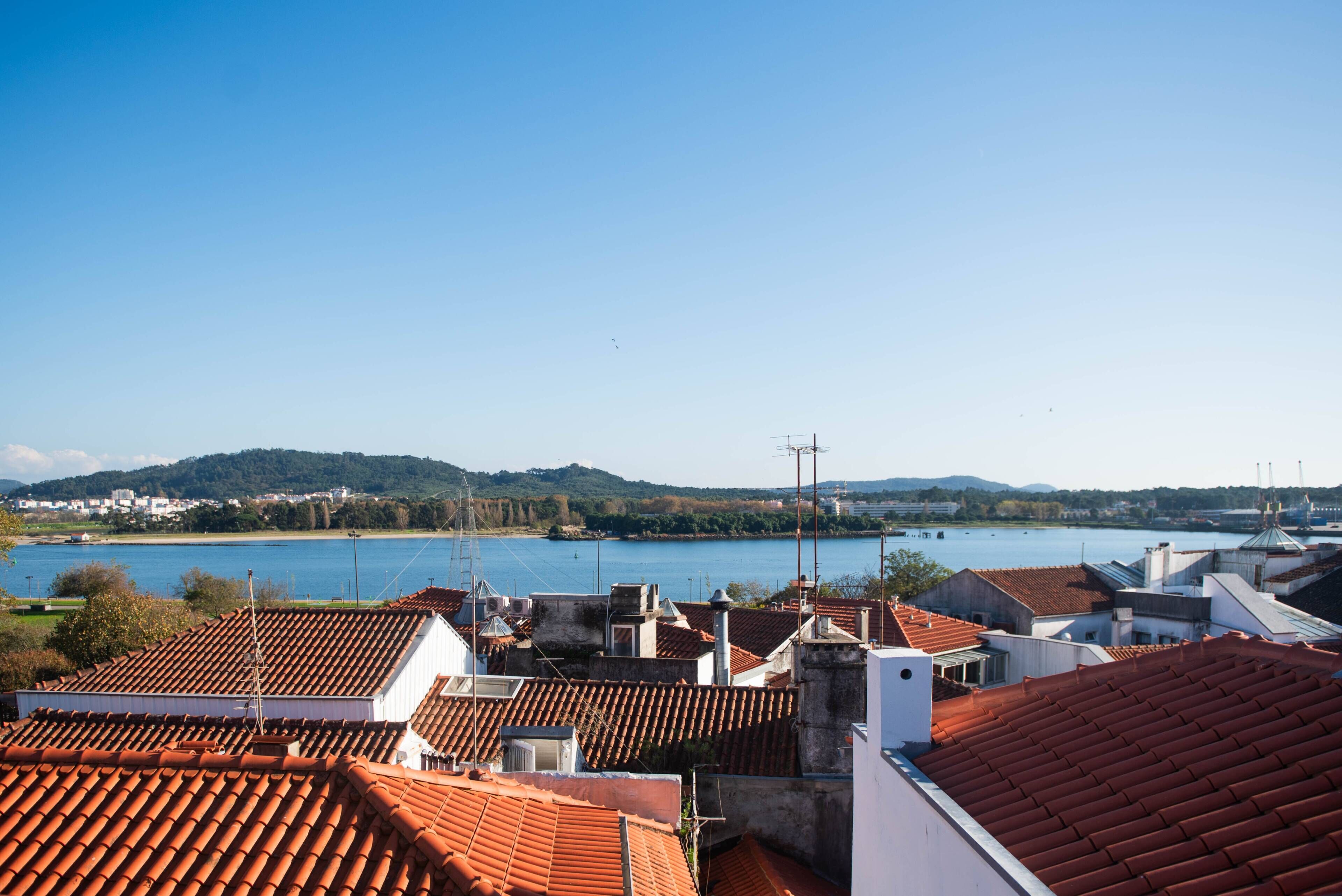 Une vue sur des toits recouverts de tuiles en terre cuite, surplombant une étendue d’eau, avec des collines et des bâtiments qui se dessinent à l’horizon.