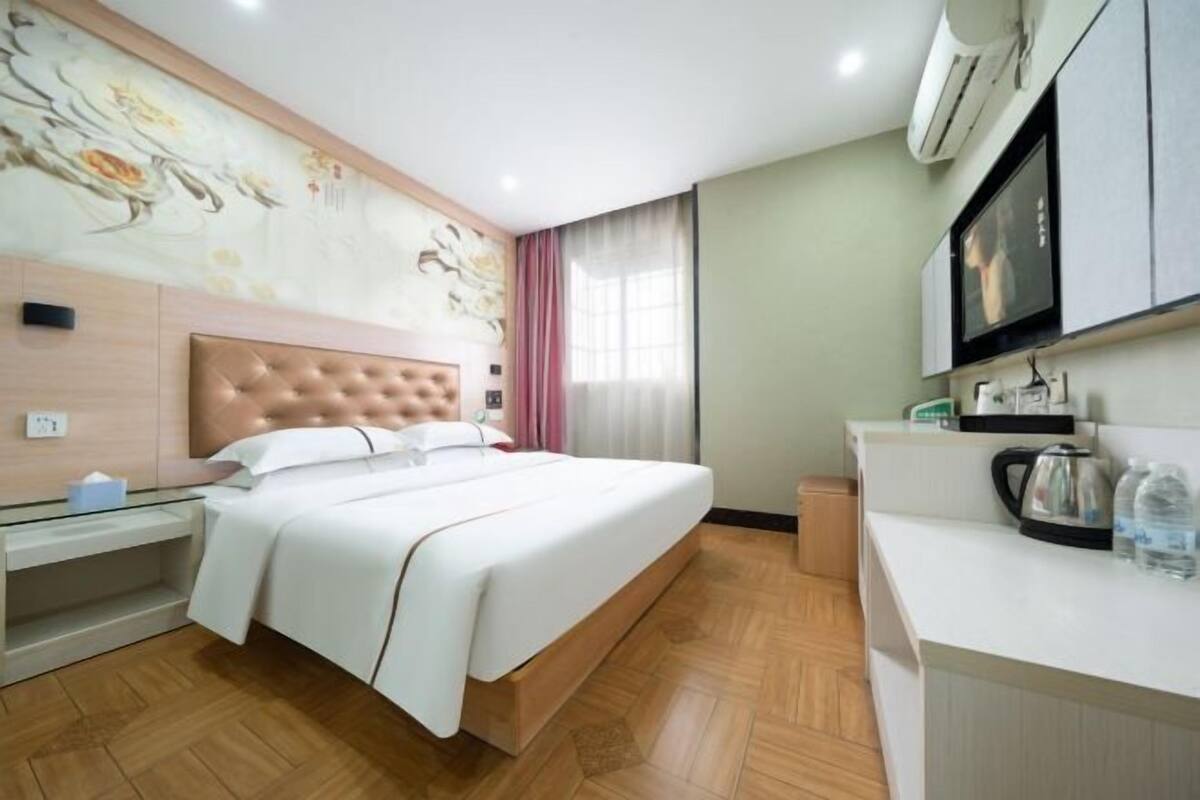 A hotel room with a large bed, a wooden headboard, a bedside table with a lamp, a flat-screen TV mounted above the bed, a small kitchenette with a kettle and a water bottle, and a decorative wall mural.