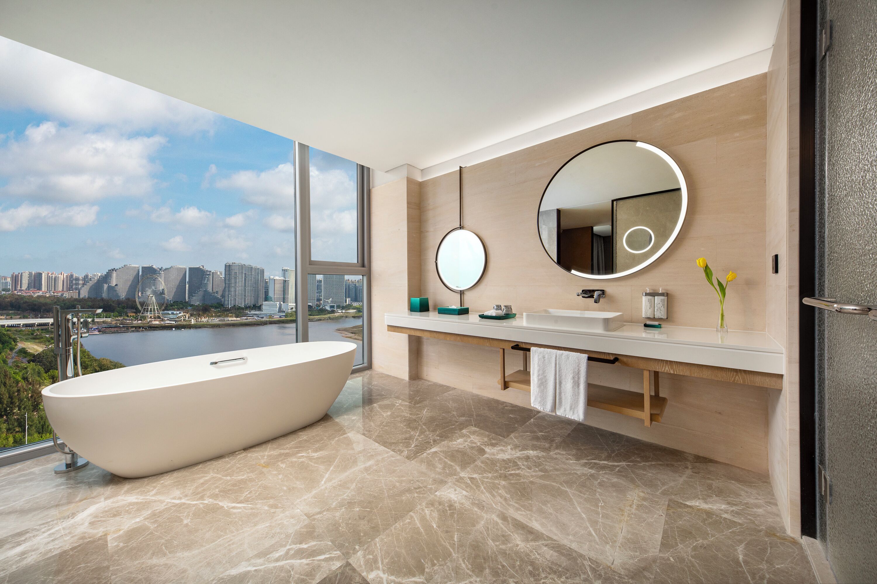 A modern bathroom with a freestanding bathtub, a large mirror, and a view of a cityscape through a large window.