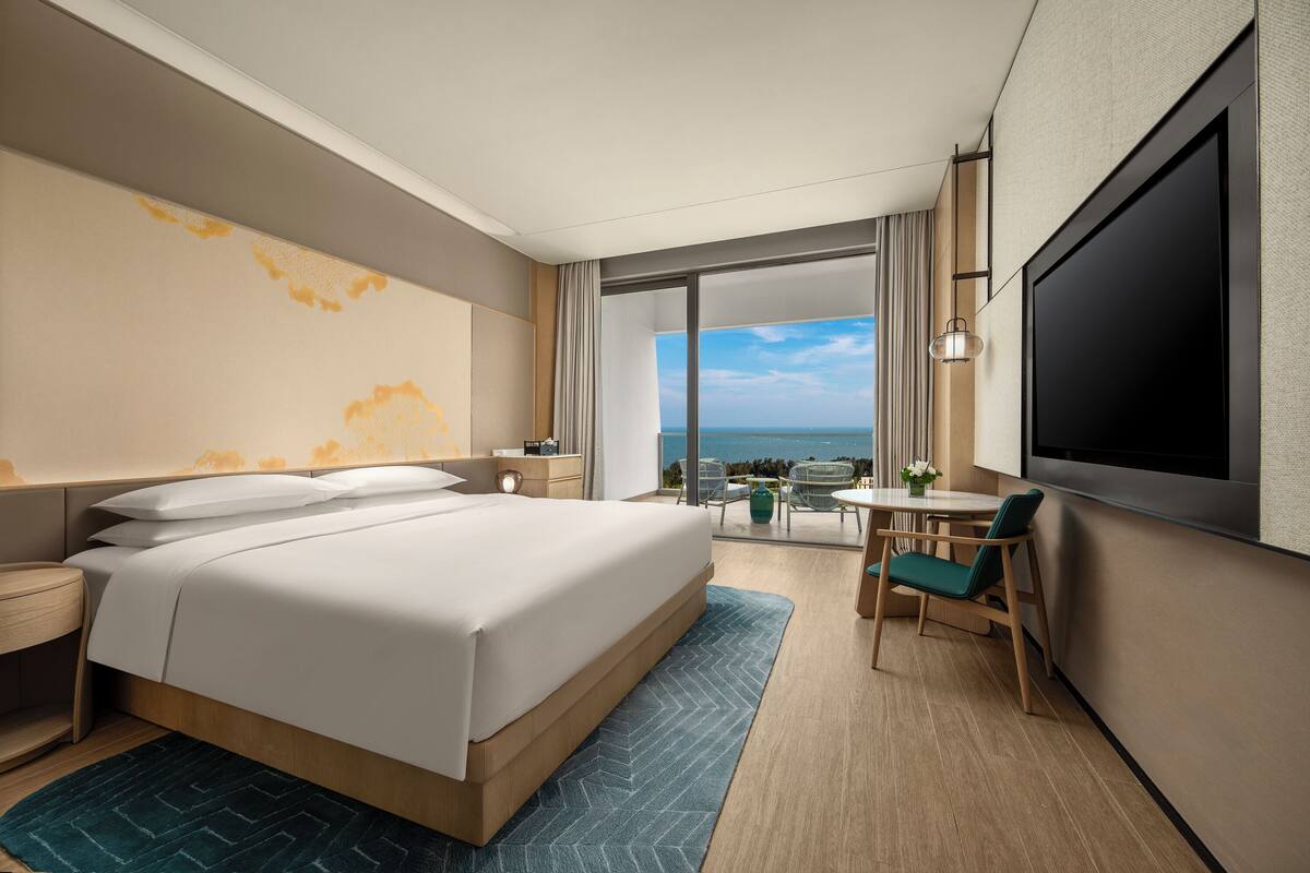 A hotel room with a large bed, a TV, a desk, and a view of the ocean.