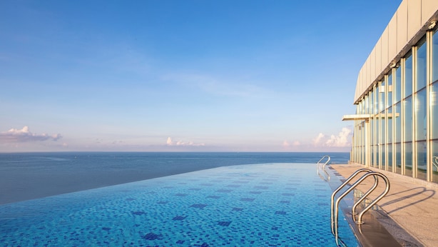 An infinity pool with a clear view of the ocean, a glass railing, and a modern building facade.