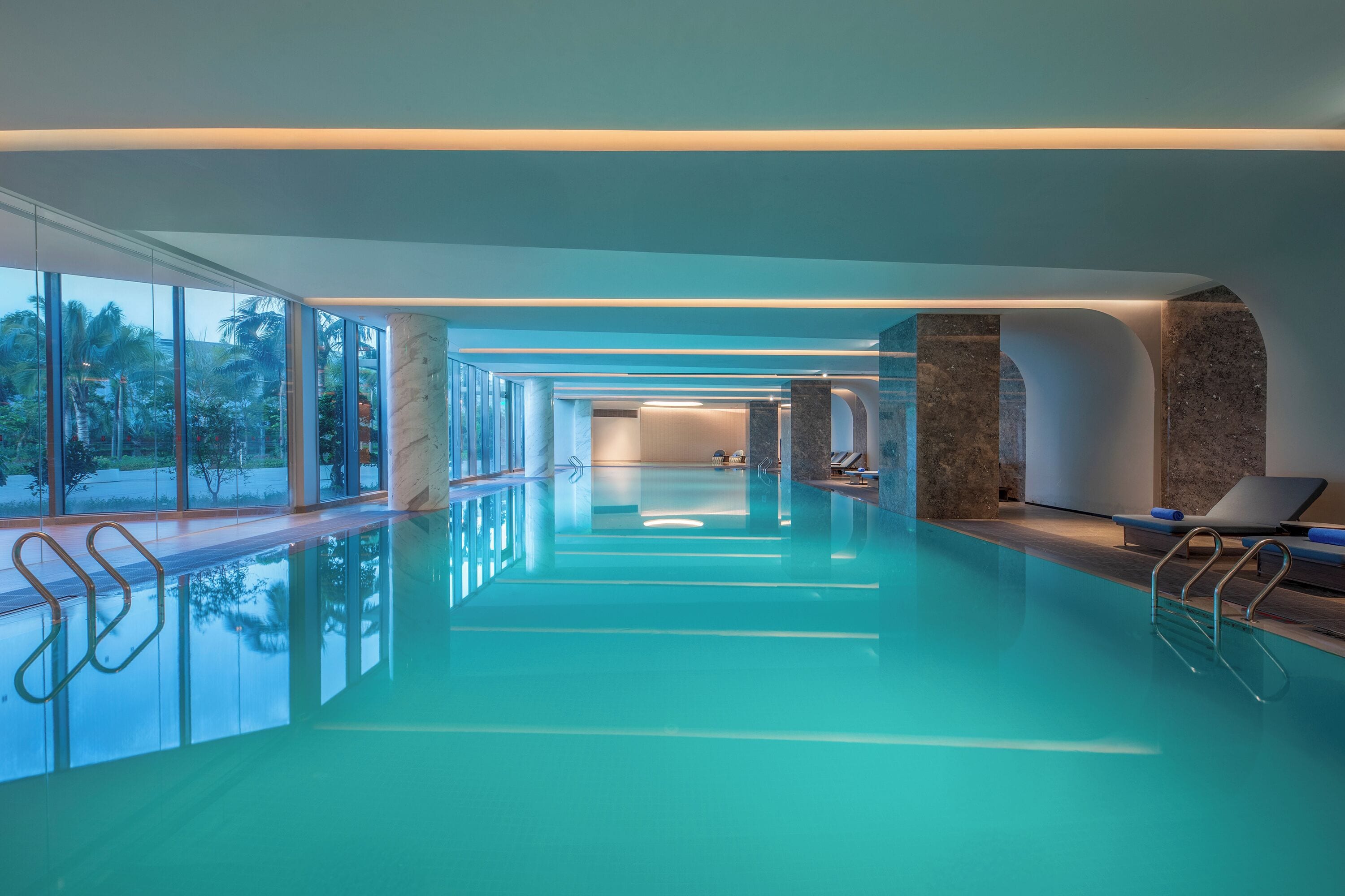 Une piscine intérieure moderne avec des parois en verre transparent, un espace détente avec des chaises longues et une vue sur l’extérieur.
