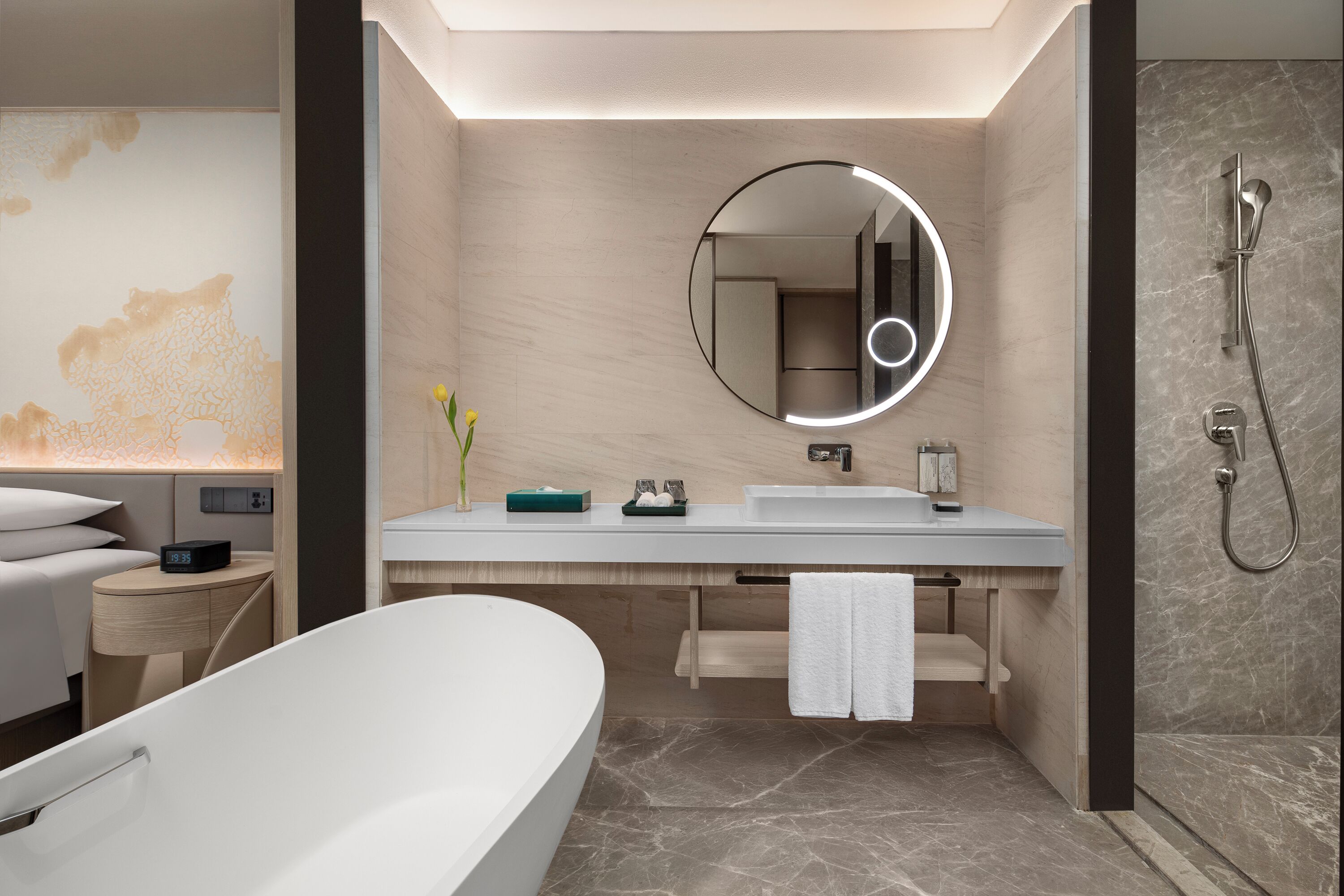 A modern bathroom with a large bathtub, a round mirror with a light, a shower, and a vanity with a sink and towel rack.