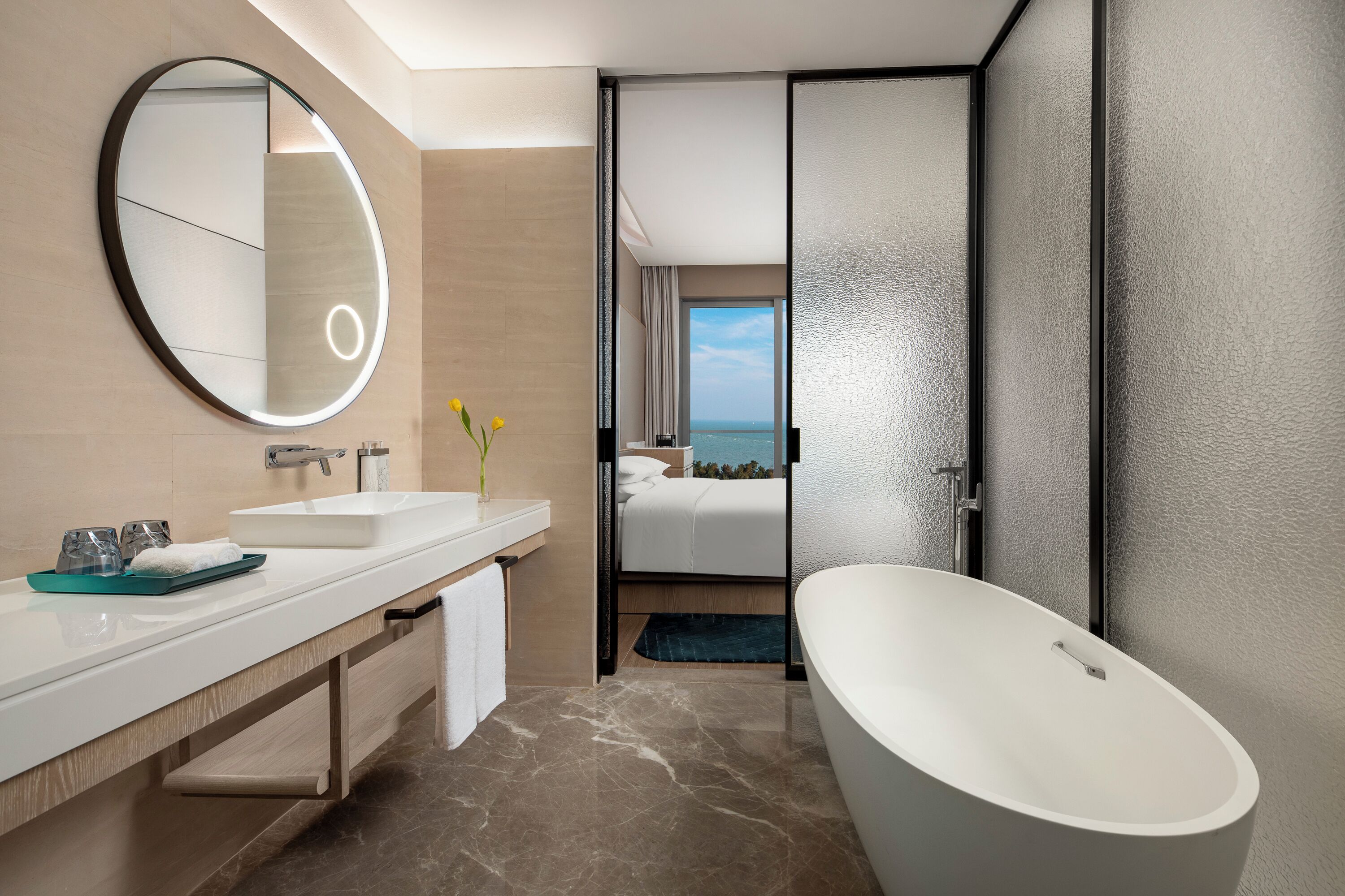 Un baño moderno con una bañera ovalada grande, un espejo redondo, un lavabo blanco y vista al mar a través de una puerta abierta.