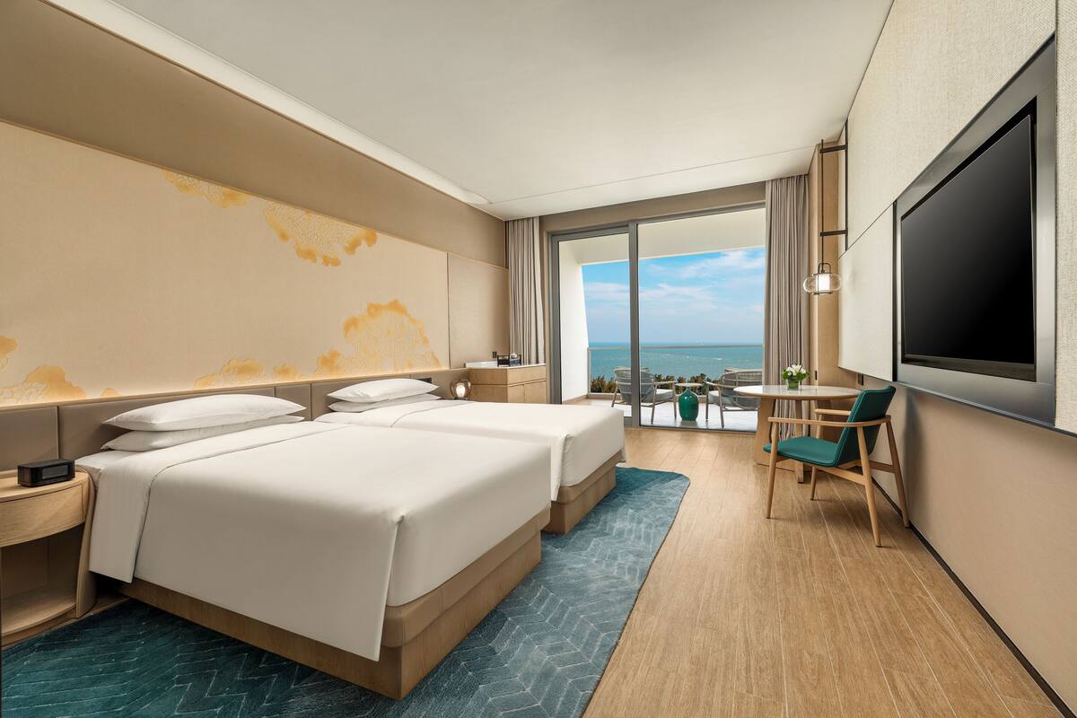A hotel room with two beds, a TV, a balcony with a view of the sea, and a small table with chairs.