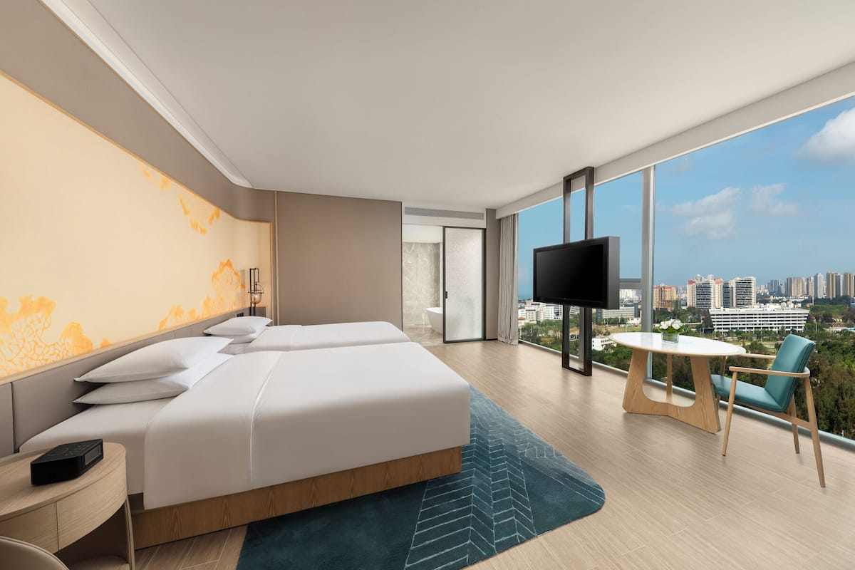 A modern hotel room with a large bed, a TV, a balcony with a table and chairs, and a city view.