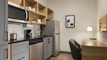 A compact kitchen with a microwave, coffee maker, and refrigerator. There is a wooden shelf above the sink holding cups and glasses. A framed picture hangs on the wall, and a desk with a lamp is visible.