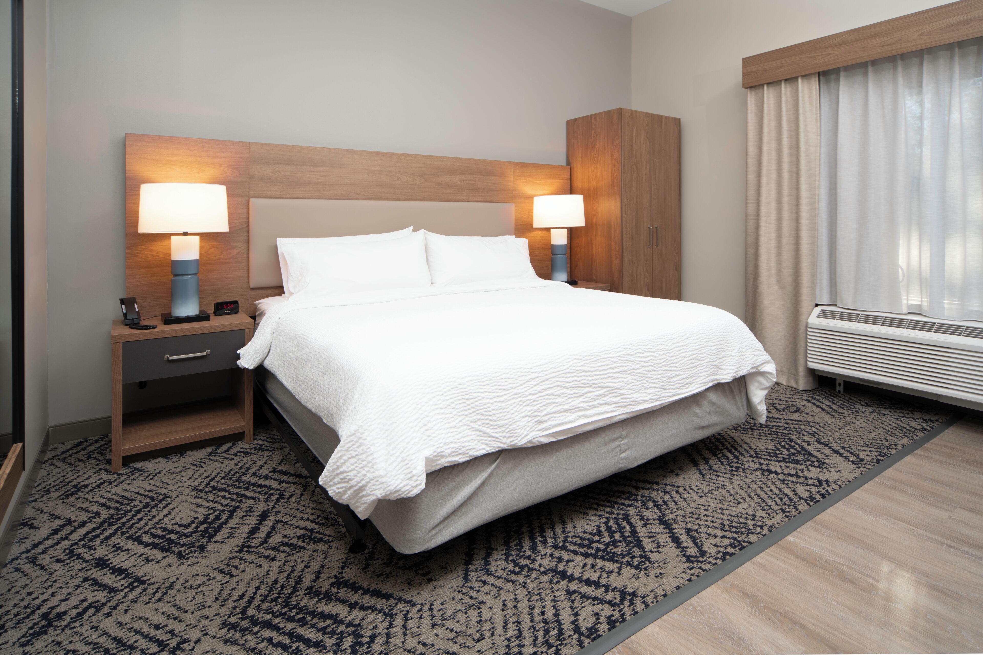 A hotel room with a large bed, two bedside tables with lamps, a nightstand, a window with curtains, and a wall-mounted heater.