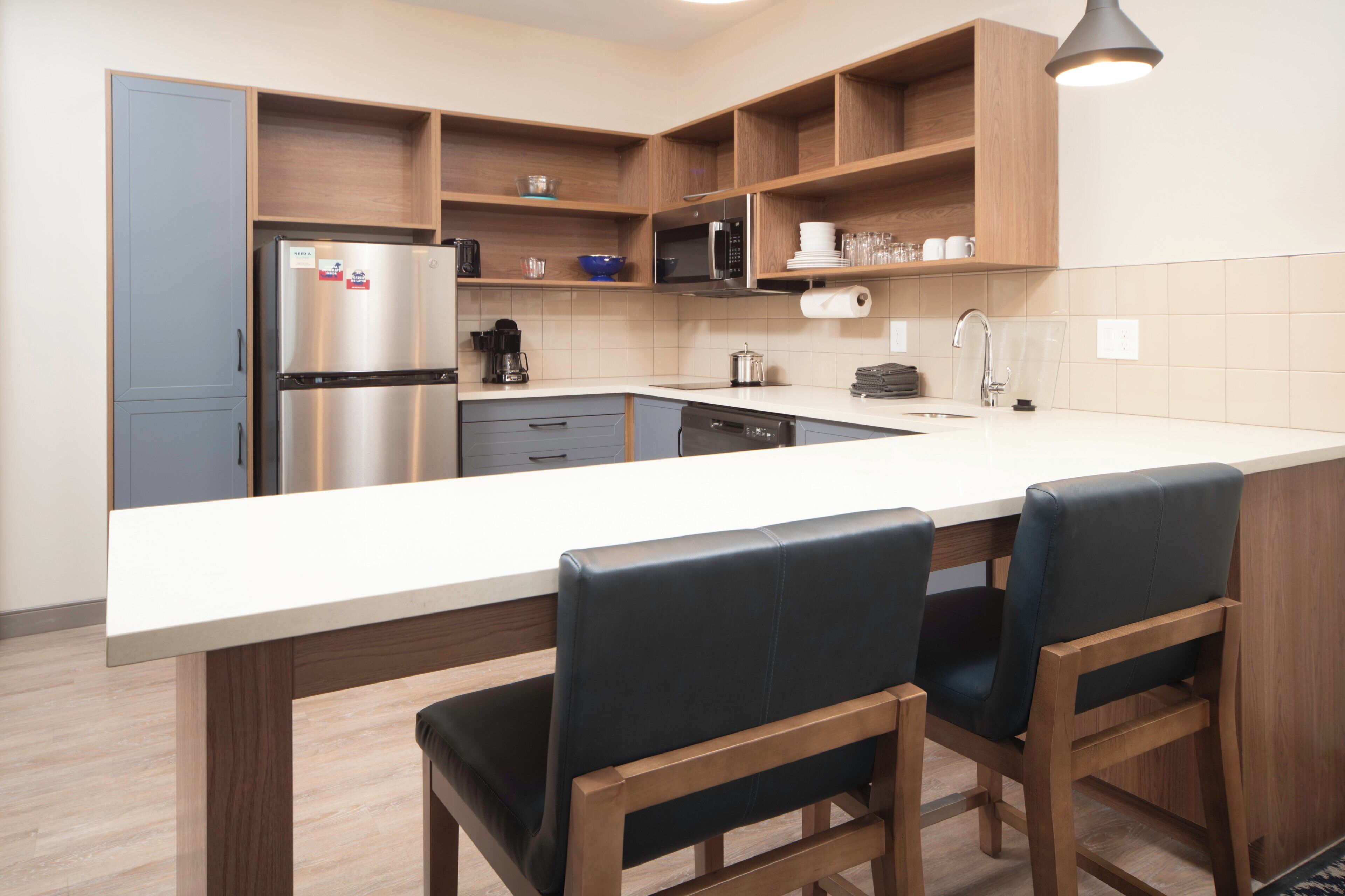 A modern kitchen with a stainless steel refrigerator, a microwave, a coffee maker, and a sink. There are wooden cabinets and a large island with bar stools.