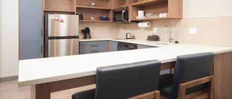 A modern kitchen with a stainless steel refrigerator, a microwave, a coffee maker, and a sink. There are wooden cabinets and a large island with bar stools.
