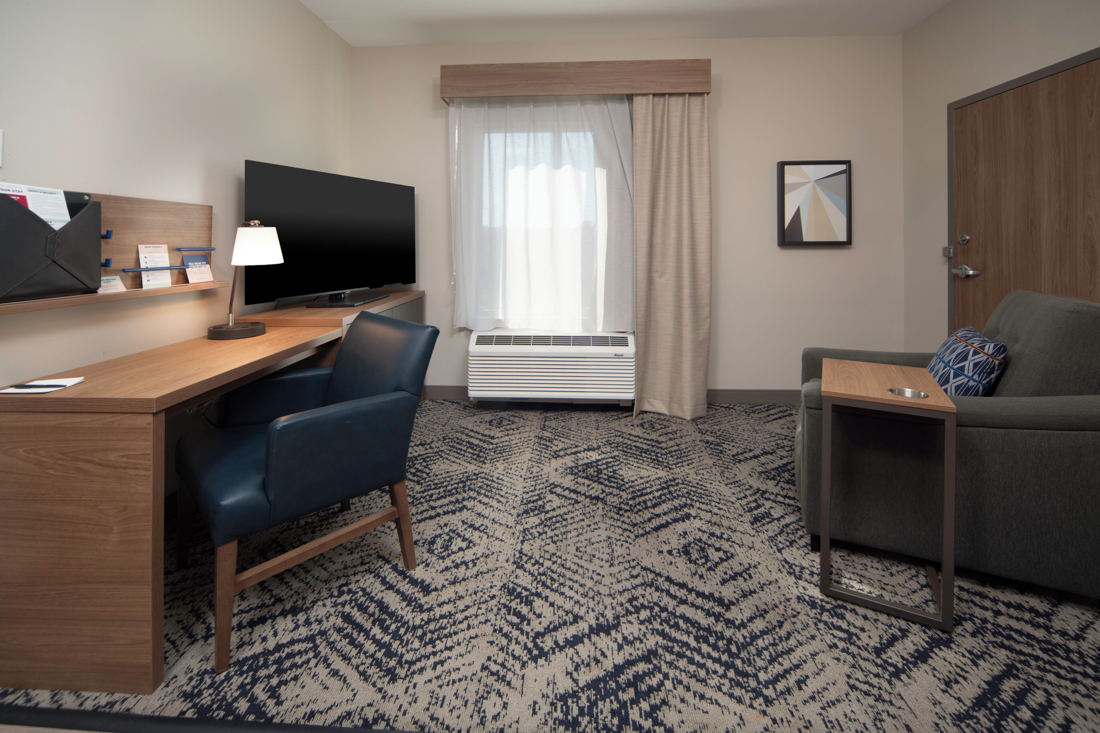 Une chambre d’hôtel équipée d’un bureau, d’une télévision, d’une chaise, d’un canapé, d’une petite table et d’une fenêtre avec des rideaux.