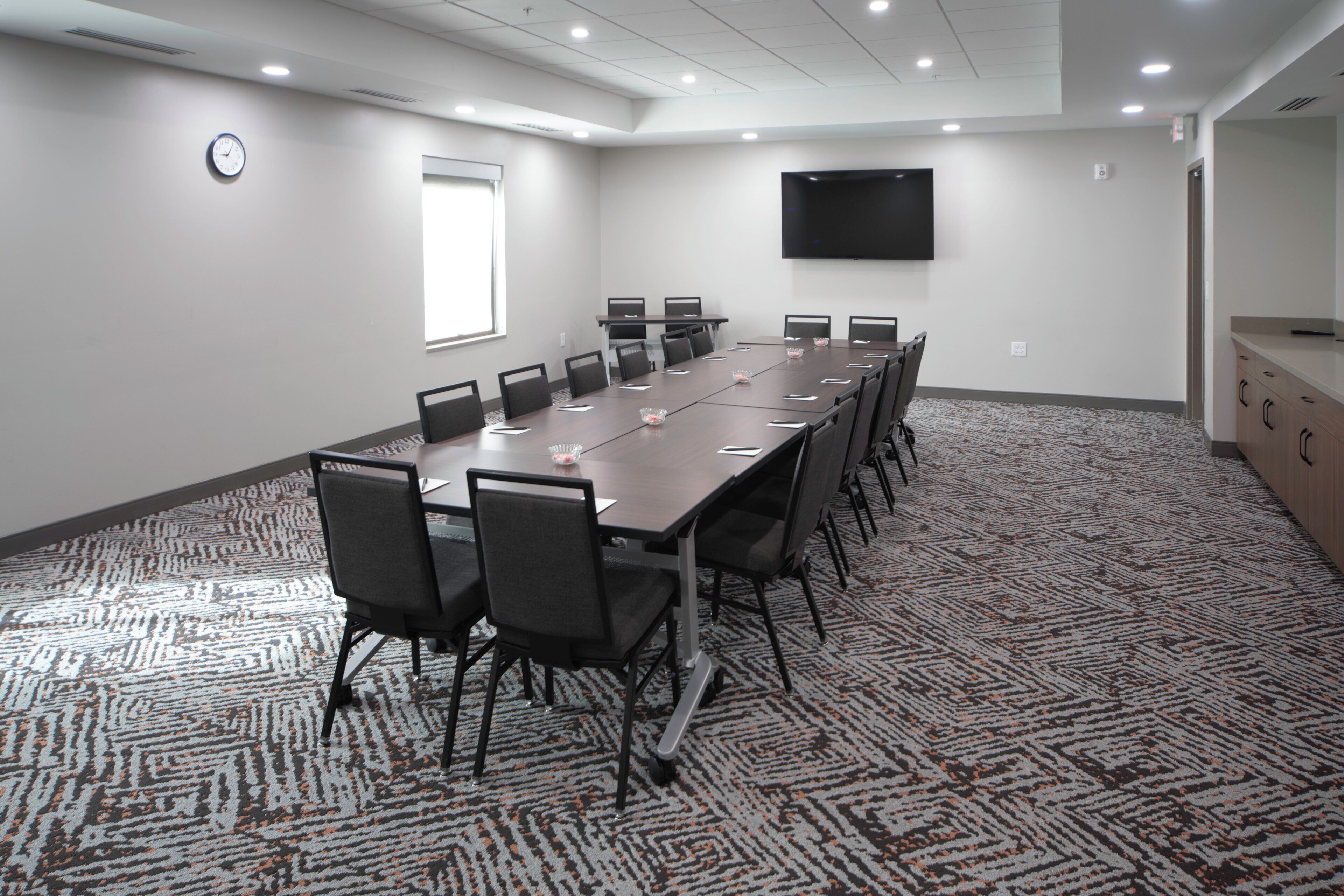 A conference room with a long table, chairs, a clock, a TV, and a patterned carpet.