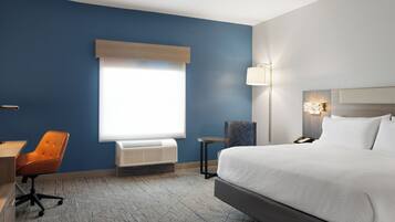 Une chambre d’hôtel avec un grand lit, un bureau, une chaise orange, une petite table, une fenêtre avec des stores et une lampe fixée au mur.