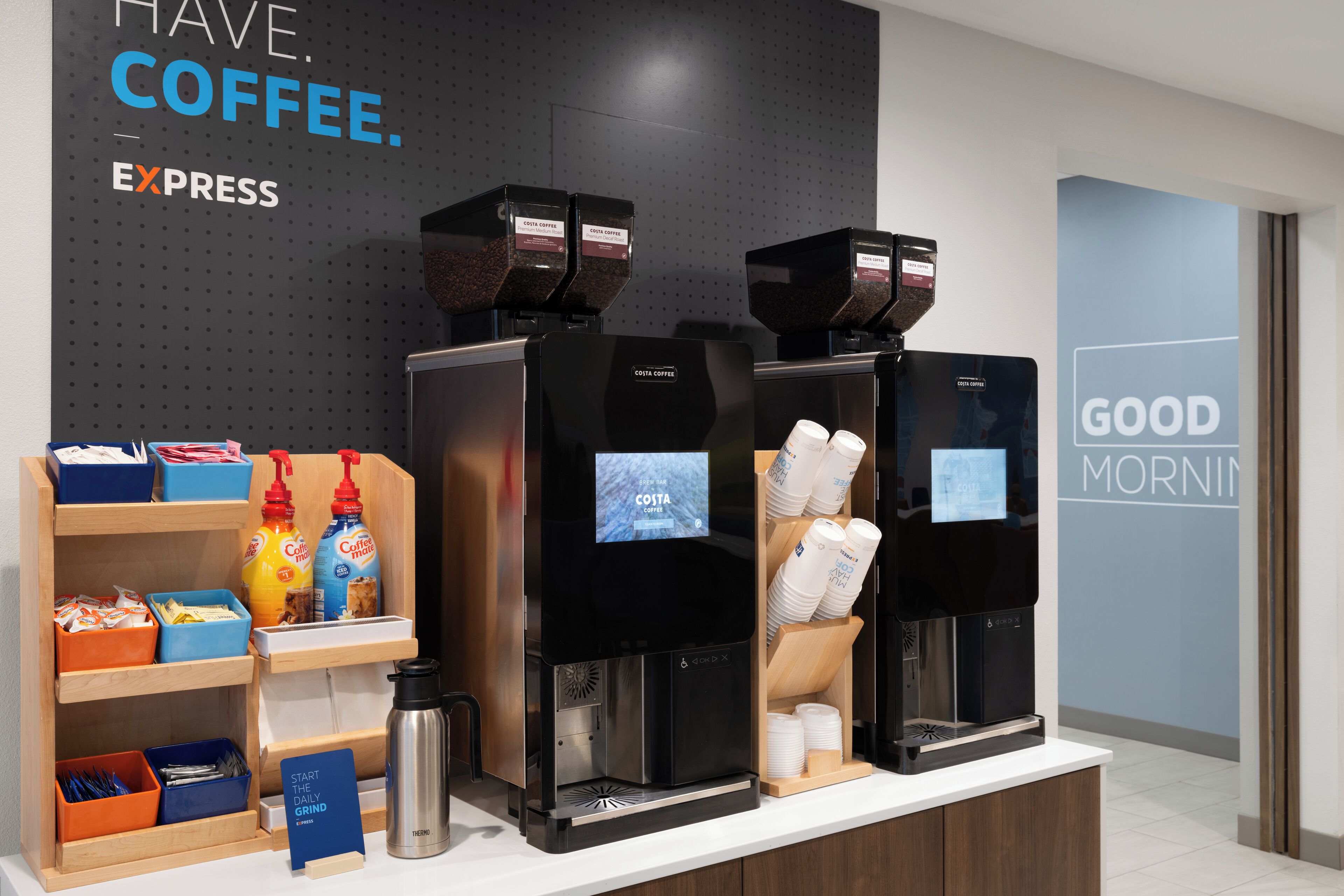 Kahvilanurkkaus, jossa on kolme espressokoneetta, ja koneiden yläpuolella on kyltti, jossa lukee: »Costa Coffee». Seinällä on myös teksti: »HANKI KAHVIA. HETI.»