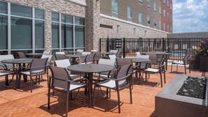 An outdoor patio area with multiple round tables and chairs, a stone wall, and a building with large windows in the background. - Holiday Inn Express & Suites Meridian I 20 by IHG (Meridian)