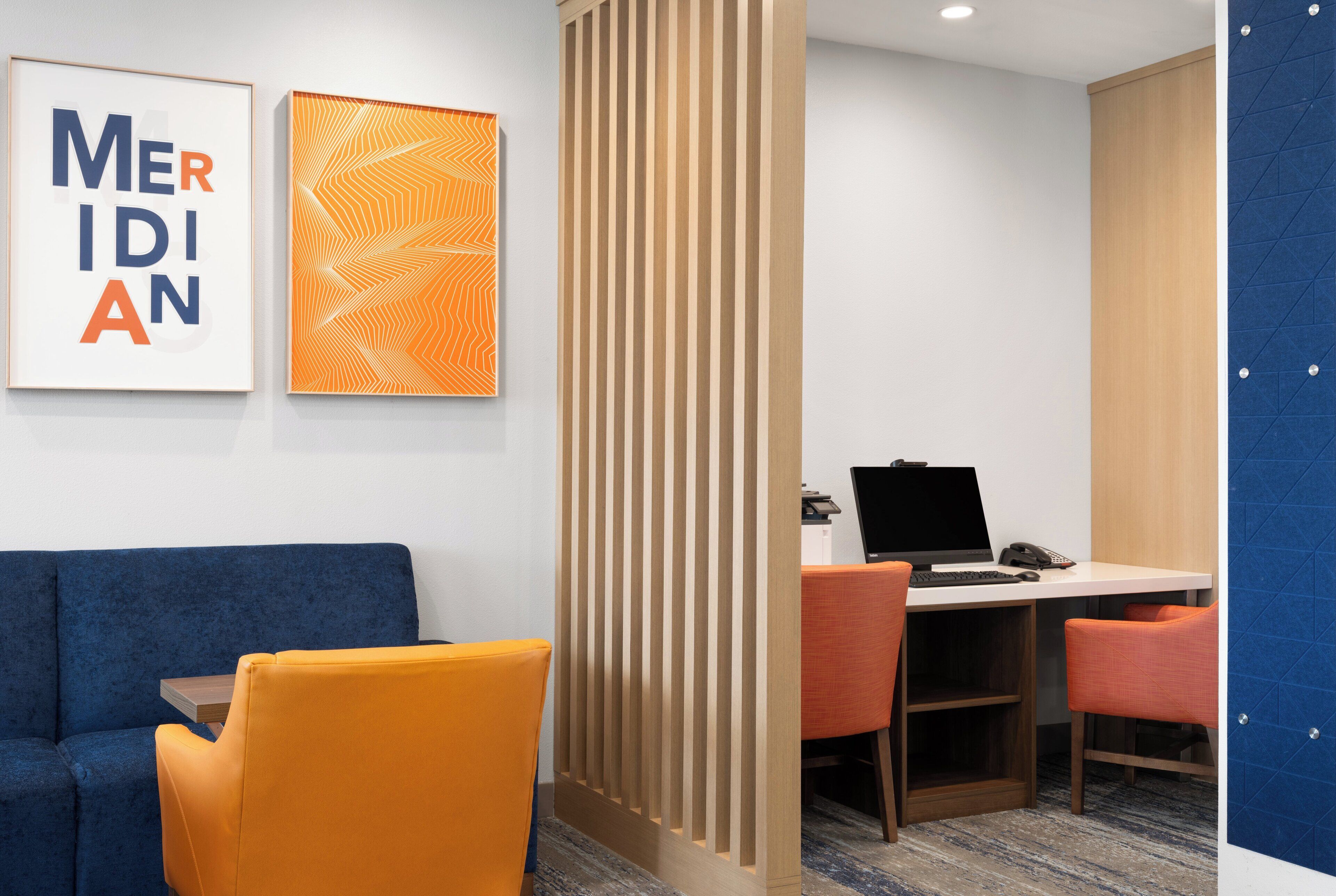 A hotel room with a blue sofa, an orange chair, a desk with a laptop, and two framed artworks on the wall.