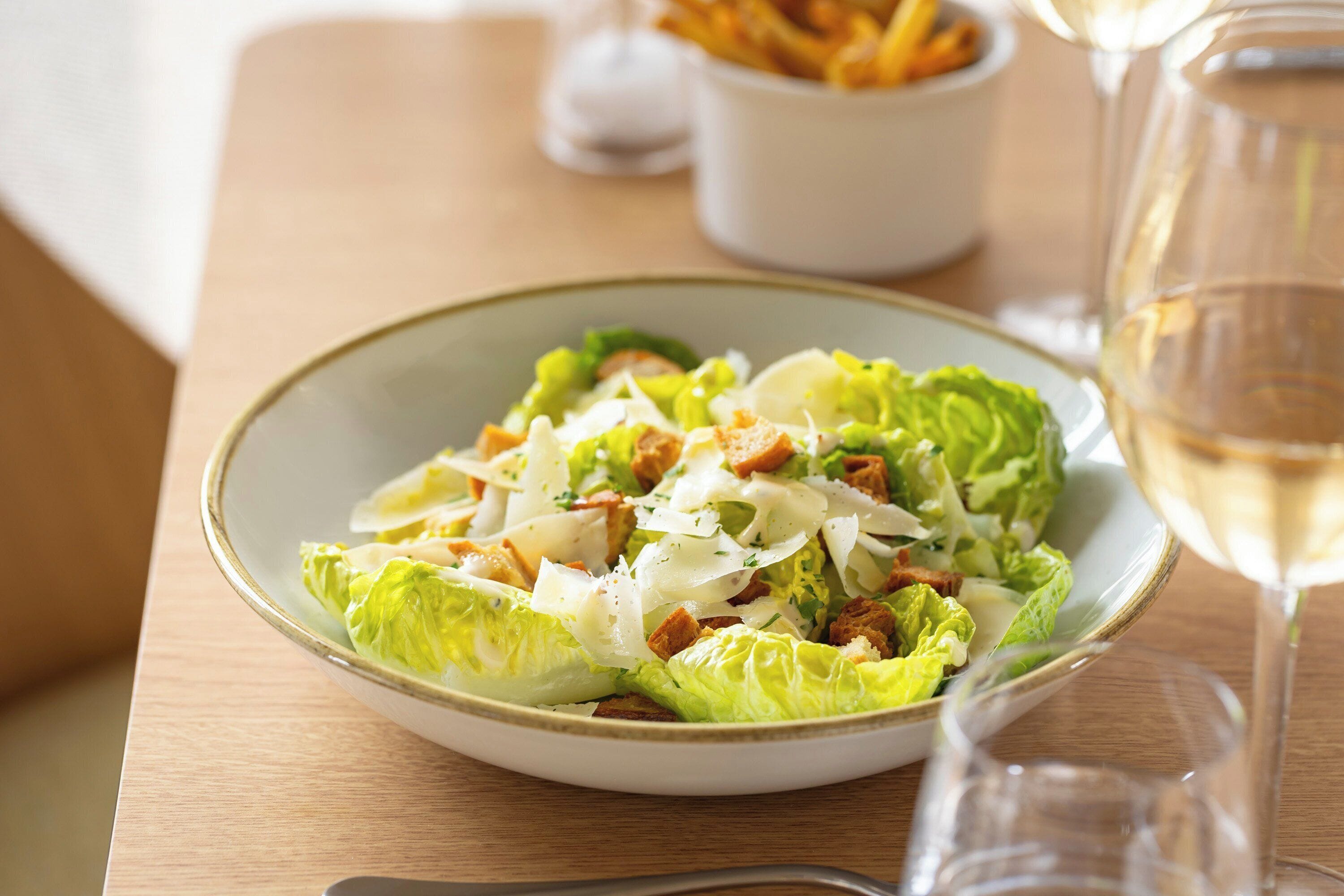 A plate of Caesar salad with croutons and cheese, a glass of white wine, and a bowl of fries on a wooden table.