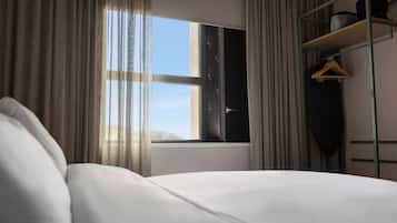 Quarto de hotel com cama, janela com cortinas, um cabideiro e uma prateleira com objetos.