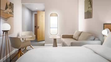 Una camera d'albergo moderna con un letto grande, un divano, un tavolino rotondo e una sedia. Si vede una porta che conduce a un'altra stanza e una finestra con vista.