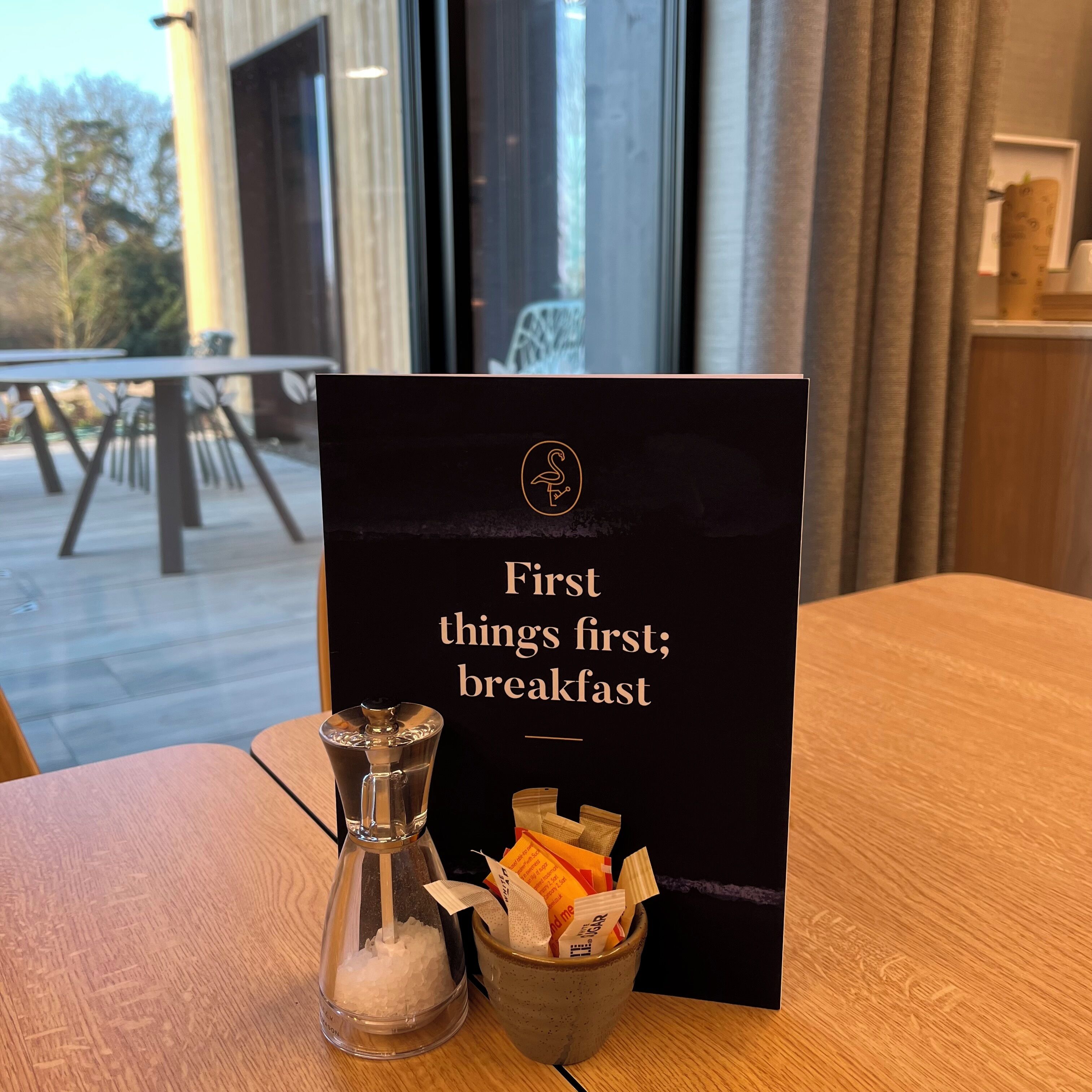 Un letrero negro con letras doradas que dice “Primero lo primero; desayuno” y un pequeño recipiente de vidrio con cubitos de azúcar y una cuchara sobre una mesa de madera.