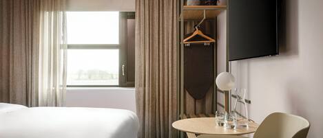 A hotel room with two beds, a small table with glasses, a hanging coat rack, and a TV mounted on the wall.