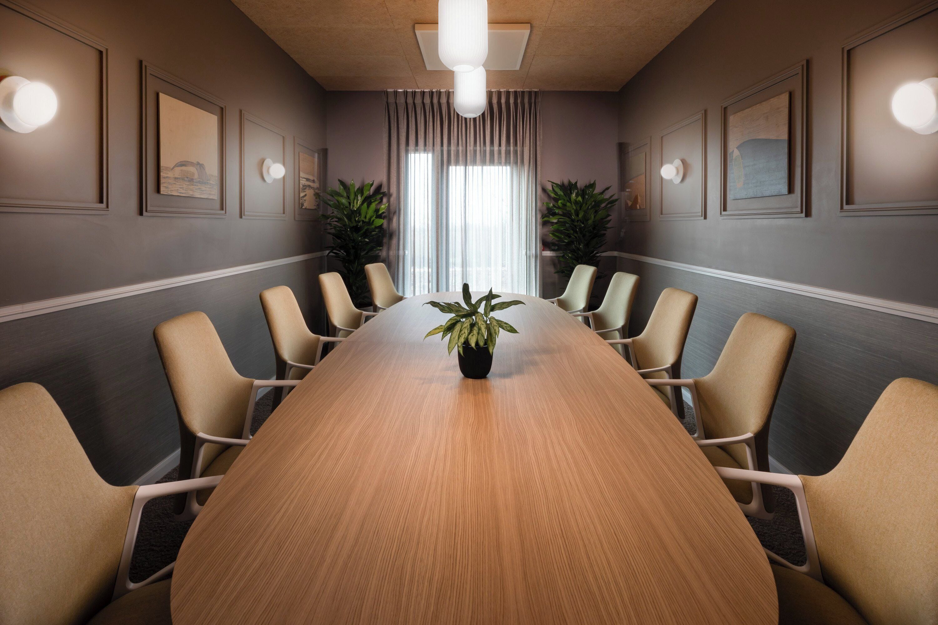 A conference room with a long wooden table, beige chairs, potted plants, framed artwork on the walls, and a large window with sheer curtains.
