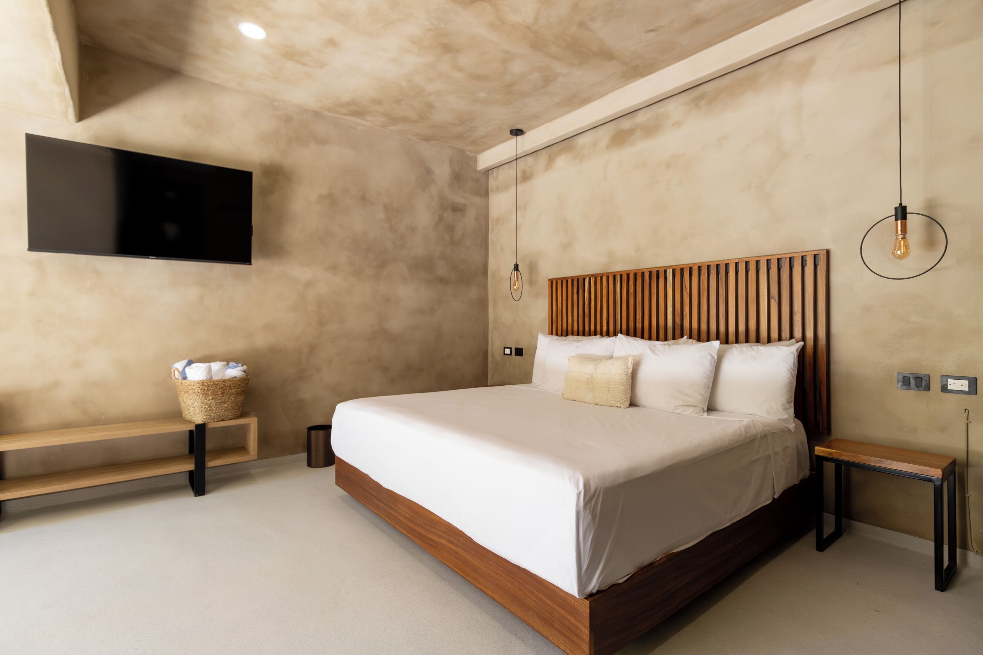 A modern bedroom with a large bed, wooden headboard, a flat-screen TV mounted on the wall, and a small bedside table with a lamp.
