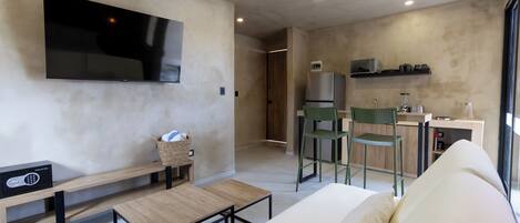 A modern living room with a flat-screen TV, a sofa, a coffee table, and a bench. There is a kitchen area with a refrigerator, microwave, and a small island with a sink and bar tools.