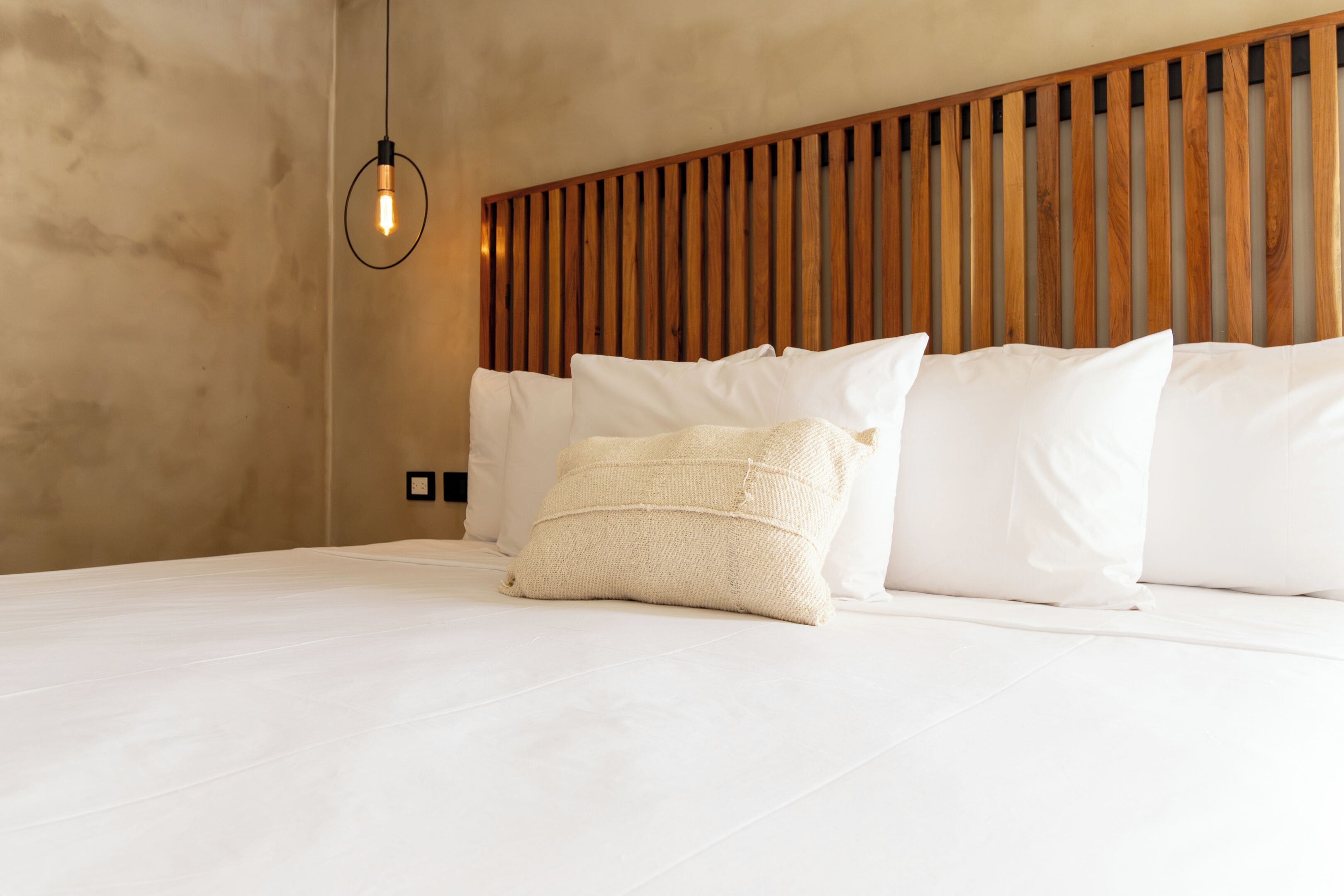 A bedroom with a wooden slat headboard, white bedding, and a single hanging light bulb.