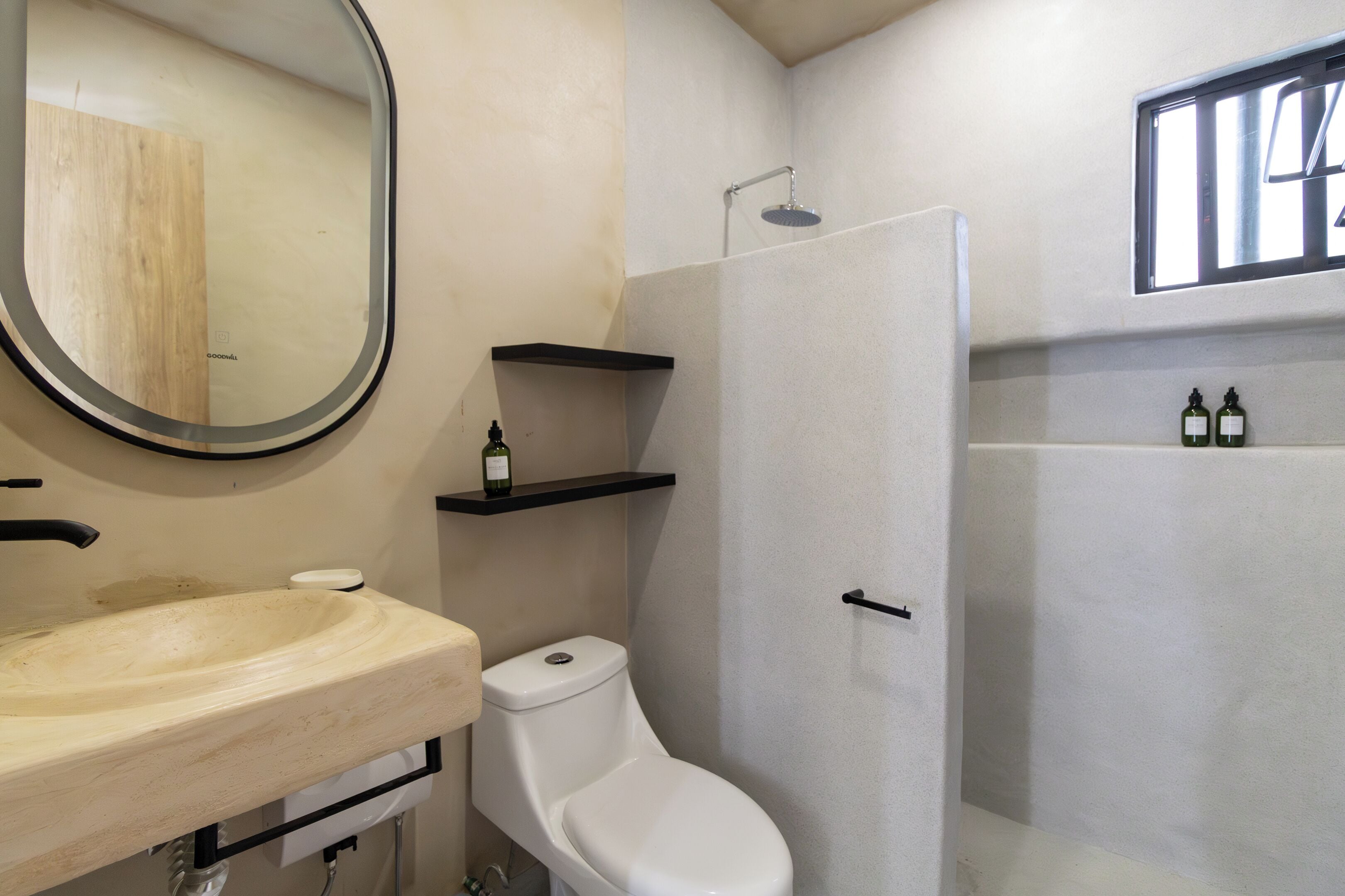 A bathroom with a sink, toilet, and shower. There are shelves with bottles and a mirror.