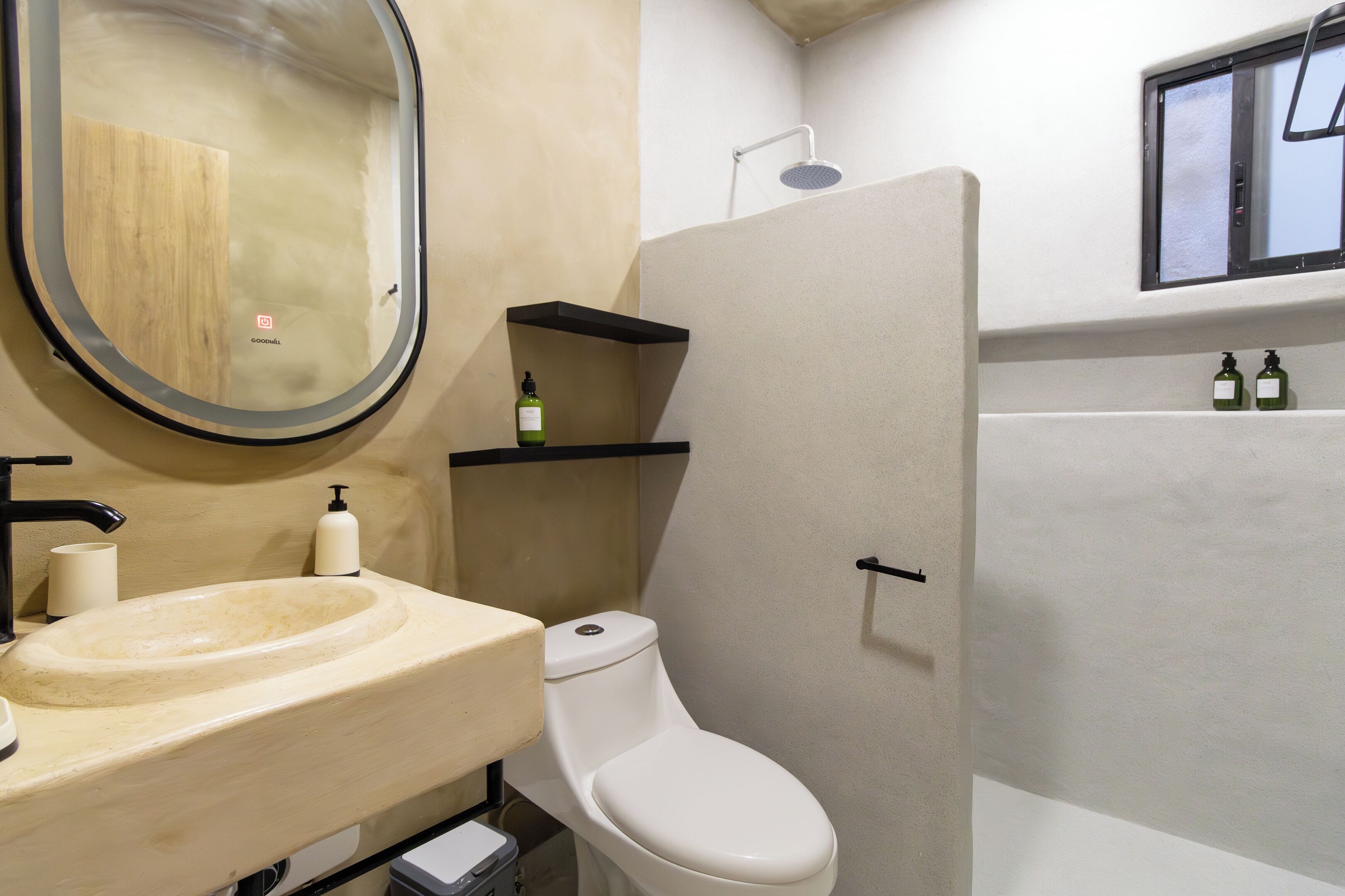 A bathroom with a beige sink, a toilet, a shower, and two shelves with bottles.