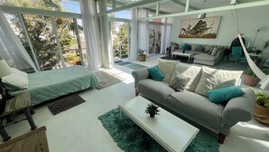 A modern living room with a bed, sofa, and coffee table. There are large windows with curtains, a hammock, and a painting on the wall. - Two story high ceiling loft with heated pool (Oaxaca de Juárez)