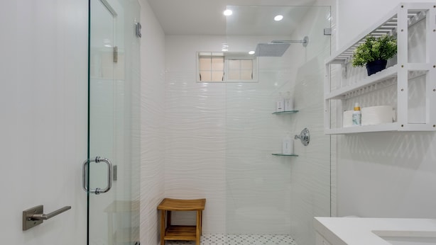Un baño moderno con cabina de ducha de vidrio, un taburete pequeño de madera, un inodoro y un estante con artículos de aseo.