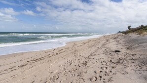 Ein sandiger Strand mit Fußspuren, die zum Wasser führen, ein klarer Himmel und ein Gebäude in der Ferne.